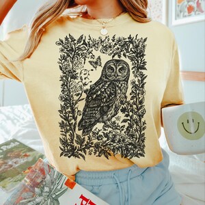 May include: A light yellow t-shirt featuring a detailed black and white illustration of an owl within a floral frame. The shirt has long sleeves and a relaxed fit. The text "Color Banana" is printed in black at the bottom right.