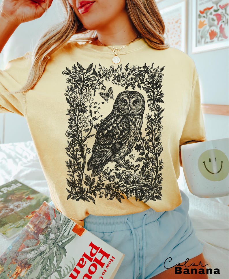 May include: A light yellow t-shirt featuring a detailed black and white illustration of an owl within a floral frame. The shirt has long sleeves and a relaxed fit. The text "Color Banana" is printed in black at the bottom right.