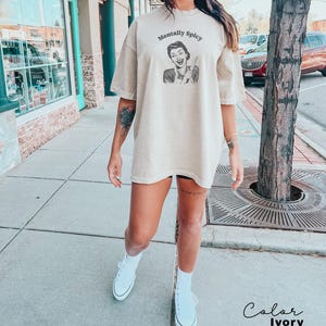 Puede incluir: Una camiseta de gran tama&ntilde;o de color beige con el texto "Mentally Spicy" y una ilustraci&oacute;n retro de una mujer sosteniendo un cuchillo. La camiseta la lleva puesta una persona con un tatuaje en el brazo. El color de la camiseta es marfil.