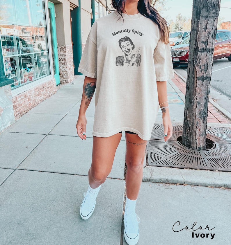 Puede incluir: Una camiseta de gran tama&ntilde;o de color beige con el texto "Mentally Spicy" y una ilustraci&oacute;n retro de una mujer sosteniendo un cuchillo. La camiseta la lleva puesta una persona con un tatuaje en el brazo. El color de la camiseta es marfil.