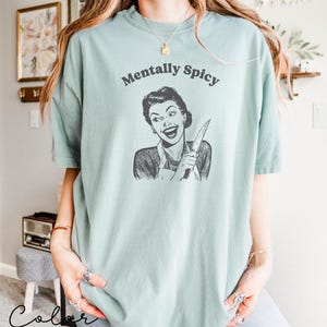 Puede incluir: Una camiseta azul claro con el texto "Mentally Spicy" impreso en la parte delantera. El texto est&aacute; en negro y el gr&aacute;fico es una ilustraci&oacute;n en blanco y negro de una mujer sosteniendo un cuchillo.