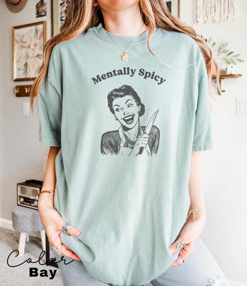 Puede incluir: Una camiseta azul claro con el texto "Mentally Spicy" impreso en la parte delantera. El texto est&aacute; en negro y el gr&aacute;fico es una ilustraci&oacute;n en blanco y negro de una mujer sosteniendo un cuchillo.