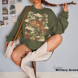 May include: Olive green sweatshirt featuring a detailed graphic of a tiger resting on a tree branch, surrounded by flowers and clouds. The design incorporates shades of brown, orange, and cream. The sweatshirt has long sleeves and a relaxed fit.