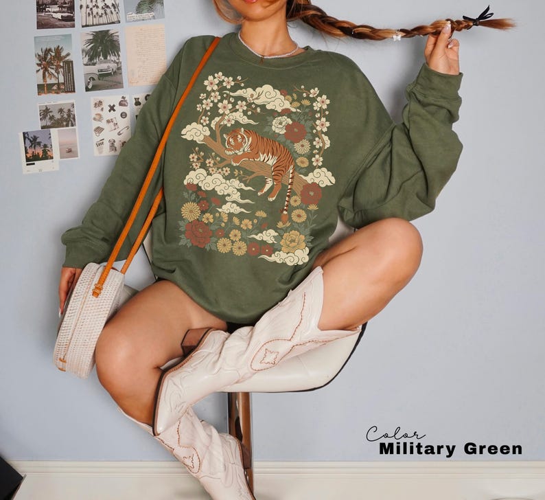 May include: Olive green sweatshirt featuring a detailed graphic of a tiger resting on a tree branch, surrounded by flowers and clouds. The design incorporates shades of brown, orange, and cream. The sweatshirt has long sleeves and a relaxed fit.