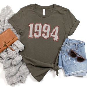 May include: A green t-shirt with the year 1994 printed in white with a red outline. The shirt is laid flat on a white background with a brown wallet, a gray sweater, and blue denim shorts.