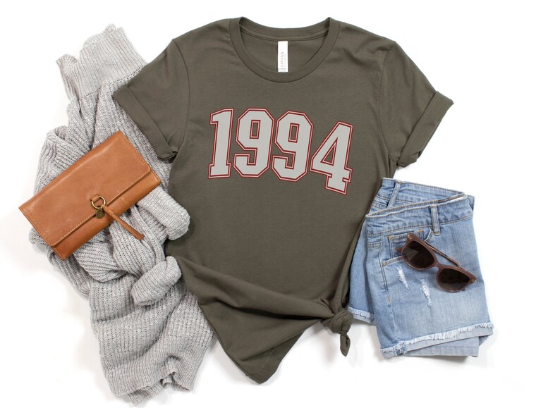 May include: A green t-shirt with the year 1994 printed in white with a red outline. The shirt is laid flat on a white background with a brown wallet, a gray sweater, and blue denim shorts.