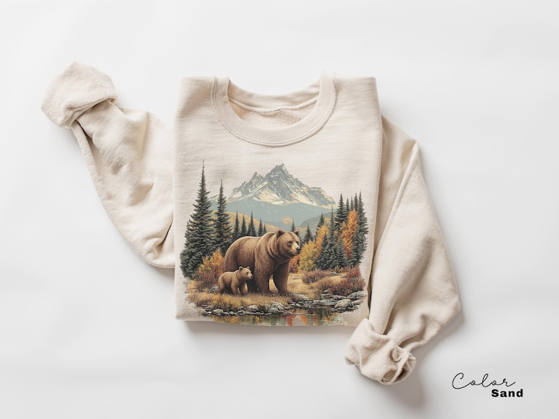 May include: A sand-colored sweatshirt featuring a detailed illustration of a bear and cub in a forest scene with a mountain backdrop. The design includes trees, a lake, and autumn foliage. The text "Color Sand" is in the bottom right corner.