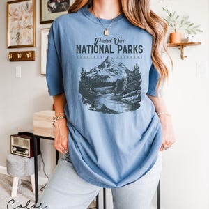 May include: A blue jean t-shirt with a graphic of a mountain range and a river. The text "Protect Our National Parks" is printed on the shirt.