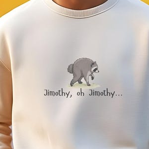 May include: A cream-colored crewneck sweatshirt featuring a small illustration of a gray raccoon and the text "Jimothy, oh Jimothy..." printed in black on the center chest. The garment has long sleeves with ribbed cuffs and a ribbed neckline.