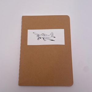 May include: A brown notebook with a simple line drawing of a small airplane on the front cover.