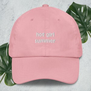 May include: A light pink baseball cap with a curved brim and an adjustable strap at the back. The front of the cap features the embroidered white text "hot girl summer" centred in a simple, clean font across the two front panels.