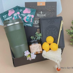 Puede incluir: Una caja de regalo que contiene un vaso de viaje verde oliva mate, un diario negro con un bol&iacute;grafo dorado, un frasco peque&ntilde;o con la etiqueta Bath Salts, tres esferas amarillas, una bolsa de malla peque&ntilde;a, dos paquetes verde azulado con la etiqueta Copper y un paquete de Rogue Jerky Co.