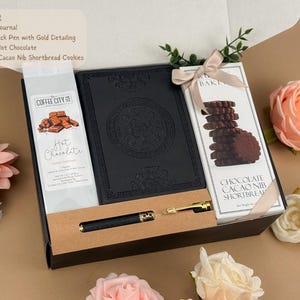 Puede incluir: Una caja de regalo que contiene un diario negro en relieve, un bol&iacute;grafo negro con detalles dorados, un paquete de chocolate caliente Coffee City y una caja de galletas de mantequilla con cacao, atada con una cinta beige en una caja negra.