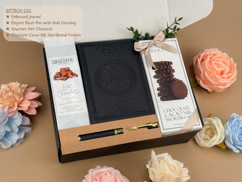 Puede incluir: Una caja de regalo que contiene un diario negro en relieve, un bol&iacute;grafo negro con detalles dorados, un paquete de chocolate caliente Coffee City y una caja de galletas de mantequilla con cacao, atada con una cinta beige en una caja negra.