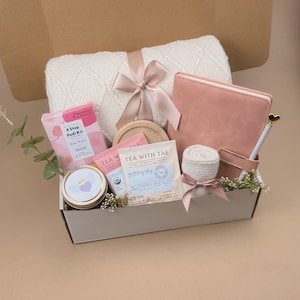 May include: A gift box containing a white knit blanket with a pink ribbon, a pink journal with a gold pen, a 4 Step Pedi Kit, a round wooden coaster, two Tea With Tae tea bags, a small candle with a heart label, and a rolled white cloth.