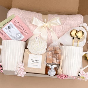 May include: A gift box filled with a pink blanket, two white ribbed mugs, tea packets, a candle, caramel candies, honey, and gold spoons. The box is tied with a cream-colored bow and decorated with pink flowers.