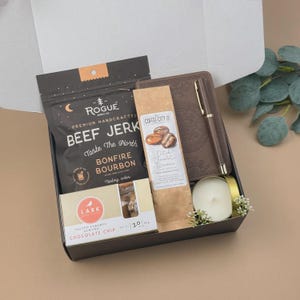 Puede incluir: Una caja de regalo que contiene una bolsa negra de carne seca Rogue, un cuaderno marr&oacute;n con un bol&iacute;grafo a juego, una bolsa de caf&eacute; Coffee City Dutch Chocolate Brownie, una caja de galletas Lark Salted Caramel Almond Chocolate Chip y una vela.