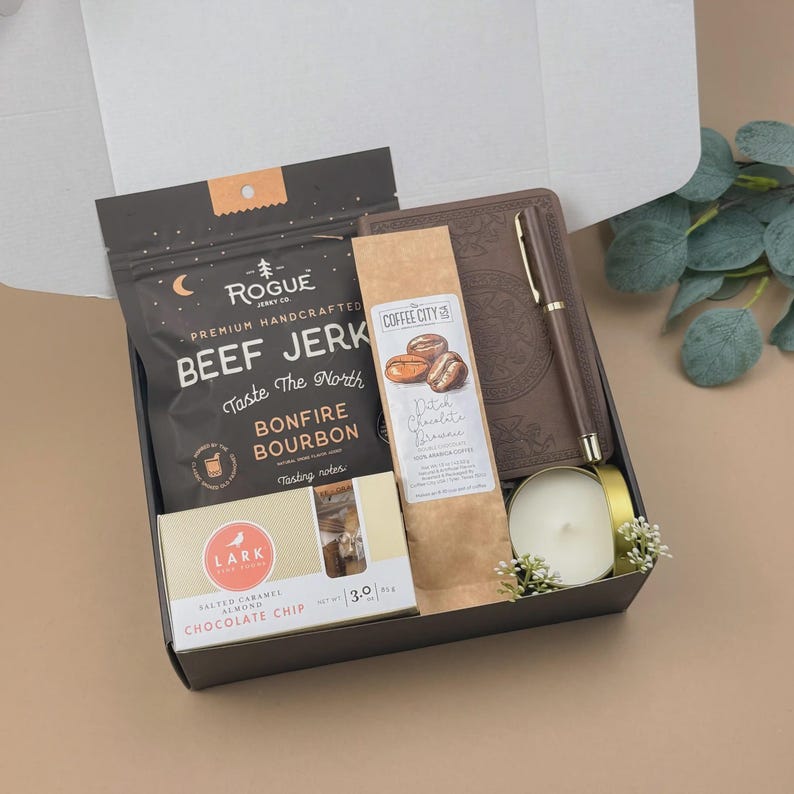 Puede incluir: Una caja de regalo que contiene una bolsa negra de carne seca Rogue, un cuaderno marr&oacute;n con un bol&iacute;grafo a juego, una bolsa de caf&eacute; Coffee City Dutch Chocolate Brownie, una caja de galletas Lark Salted Caramel Almond Chocolate Chip y una vela.