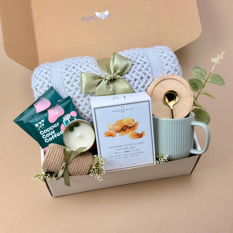 Puede incluir: Una caja de regalo que contiene una manta de punto trenzado gris claro con una cinta verde salvia, dos paquetes de caf&eacute; Copper Cow, una vela peque&ntilde;a, una caja de patatas fritas artesanales Jocelyn & Co. y una taza verde salvia acanalada.