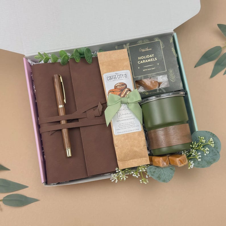 Puede incluir: Una caja de regalo que contiene un diario de cuero marr&oacute;n con un bol&iacute;grafo a juego, una bolsa de brownies de chocolate Coffee City USA con un lazo verde, una caja de caramelos navide&ntilde;os Grand Provisions y una taza verde con banda de madera.