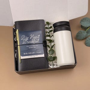 Puede incluir: Una caja de regalo que contiene un cuaderno negro con un bol&iacute;grafo dorado, un paquete de t&eacute; negro Big Heart Breakfast, una taza de viaje blanca con tapa negra y una rama decorativa de hojas verdes sobre relleno de papel blanco.
