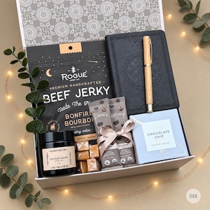 May include: A gift box containing a black bag of Rogue Jerky Co. beef jerky, a black journal with a light wood pen, two packets of Coffree coffee, a small box of chocolate chip cookies, a black jar candle, and several square caramel candies.