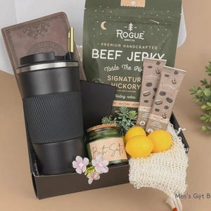 Puede incluir: Una caja de regalo que contiene un vaso de viaje negro, un cuaderno marr&oacute;n, una bolsa de carne seca Rogue, dos palitos de Coffree, un frasco de sales de ba&ntilde;o, tres limones amarillos, una esponja de malla y flores rosas. El texto Men's Gift Box 18 es visible.
