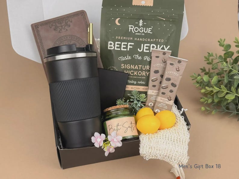 Puede incluir: Una caja de regalo que contiene un vaso de viaje negro, un cuaderno marr&oacute;n, una bolsa de carne seca Rogue, dos palitos de Coffree, un frasco de sales de ba&ntilde;o, tres limones amarillos, una esponja de malla y flores rosas. El texto Men's Gift Box 18 es visible.
