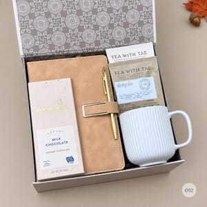May include: A gift box containing a tan notebook with a gold pen, a white ribbed ceramic mug, a packet of Tea With Tae Bombay Chai tea, and a 3-ounce bar of Moonstruck creamy milk chocolate. The box lid features a gray and white geometric pattern.
