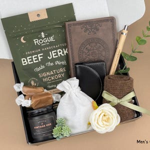 May include: A gift box filled with various items, including beef jerky, a journal, a wallet, a candle, and wrapped candies. The box also contains a pen, a small towel, a white flower, and a small bag. The text "Men's Gift Box 12" is visible.