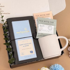 Puede incluir: Una caja de regalo que contiene un cuaderno de cuero negro, una barra de chocolate negro con almendras Moonstruck azul claro, una taza de cer&aacute;mica blanca acanalada y dos bolsitas de t&eacute; Tea With Tae. La caja incluye una lista llamada GIFTBOX-070.