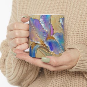May include: A white ceramic mug with a colorful iridescent blue and purple iris flower design. The background is a light beige with gold accents.