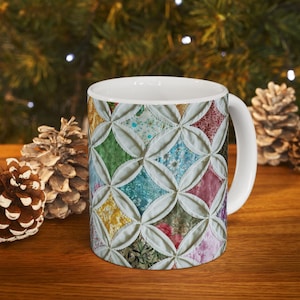 May include: White ceramic coffee mug with a colorful patchwork quilt design. The quilt pattern features diamonds in various colors, including yellow, green, blue, and pink, with white stitching. The mug has a white handle and is set on a wooden surface.