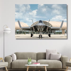 Può includere: Un aereo da caccia grigio con un abitacolo monopposto è parcheggiato su una pista di fronte a un grande edificio. L'aereo ha un design angolare distintivo con un muso appuntito e ali a freccia. Il cielo è blu con nuvole bianche.