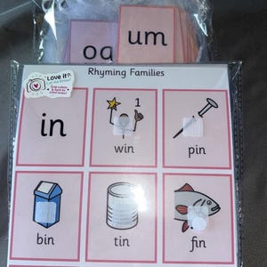 Può includere: Un set di flashcard educative con rime in una confezione di plastica trasparente. Le carte presentano parole come "in", "win" e "pin" con illustrazioni corrispondenti. È inclusa anche una carta rosa con la scritta "Grazie".