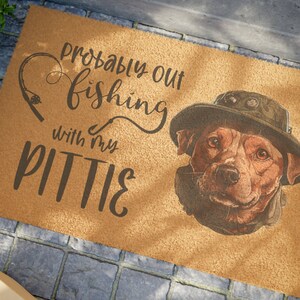 Peut inclure: Un paillasson en fibre de coco brun avec le texte "Probably out fishing with my Pittie" et une image de dessin animé d'un chien brun et blanc portant un chapeau.
