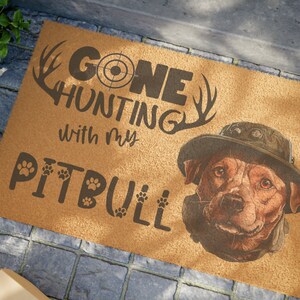 Peut inclure: Un paillasson en fibre de coco brun avec le texte "Gone Hunting with my Pitbull" en lettres noires. Le texte est entouré d'une paire de bois de cerf et d'un symbole de cible. Un pitbull brun et blanc portant un chapeau de camouflage est représenté dans le coin inférieur droit.