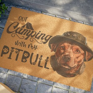 Peut inclure: Un paillasson en fibre de coco brun avec le texte "Out Camping with my Pitbull" en lettres brunes. Le texte est entouré d'empreintes de pattes. Un pitbull brun et noir portant un chapeau brun se trouve dans le coin inférieur droit du paillasson.