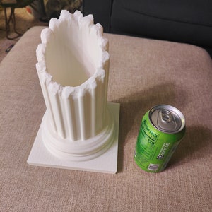 May include: White ceramic column with a square base, resembling a miniature Greek column. A green aluminum can with a nutrition facts label is next to the column.