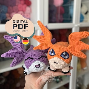 May include: A digital PDF crochet pattern featuring two plush creatures. One is purple with large wings and a white face, the other is orange with large wings and a white face. Both have black and white eyes and small black feet. Text reads DIGITAL PDF.