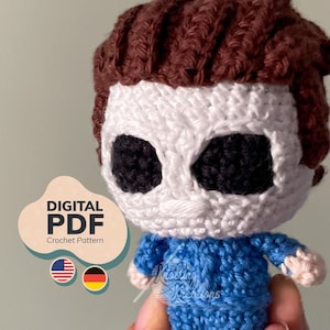 May include: A small crocheted doll featuring a white mask with large black eye sockets, brown hair, and a blue jumpsuit. A digital graphic overlay on the left side displays the text DIGITAL PDF Crochet Pattern with American and German flag icons.