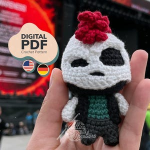 May include: A small crocheted doll featuring a white head with a red mohawk, black eye patches, and a black vest over a green shirt. The doll has white arms and black legs. A digital PDF crochet pattern label is visible in the upper left corner.