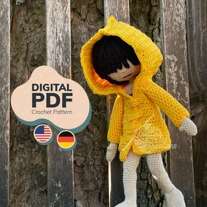 Könnte beinhalten: Eine gehäkelte Puppe mit einem leuchtend gelben Regenmantel mit Kapuze, schwarzem Haar und hellen Gliedmaßen. Das Bild enthält den Text DIGITAL PDF Crochet Pattern sowie Symbole der amerikanischen und deutschen Flagge.