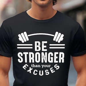 May include: Black t-shirt with the text "BE STRONGER than your EXCUSES" in white. The design includes a barbell graphic above the text. The shirt is worn by a person in an outdoor setting.