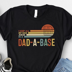 May include: Black t-shirt with the text "I KEEP ALL MY DAD JOKES IN A DAD-A-BASE" in a retro design. The graphic includes a floppy disk and horizontal stripes in orange, yellow, and blue. The shirt is paired with sneakers and jeans.