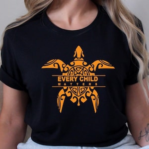 May include: Black t-shirt featuring a stylized orange turtle design with the text "EVERY CHILD MATTERS". The turtle design is centered on the shirt and has intricate patterns. The shirt is a crew neck and has short sleeves.