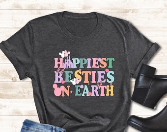 Op de afbeelding: Een donkergrijs T-shirt met de tekst "Happiest Besties on Earth" in kleurrijke letters. De tekst wordt omringd door kleine cartoonafbeeldingen van Mickey Mouse-oren, harten en een kasteel.