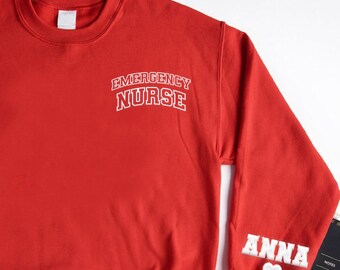 May include: A red sweatshirt with the text "EMERGENCY NURSE" in white lettering on the chest. The right sleeve has the name "ANNA" in white lettering with a white heart. Other items include a watch, glasses, a wallet, and a notebook.