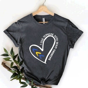 May include: A dark gray t-shirt with a white heart design. The heart has yellow and blue accents and the text "I love someone with Down Syndrome" is written around the heart.