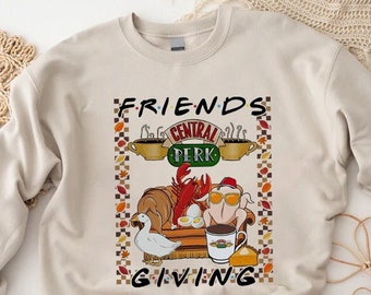 Puede incluir: Sudadera beige con un gráfico que presenta el logotipo de Central Perk y las palabras "Friends Giving". El diseño incluye un pavo con gafas de sol, una langosta, un ganso y tazas de café. La sudadera tiene mangas largas.